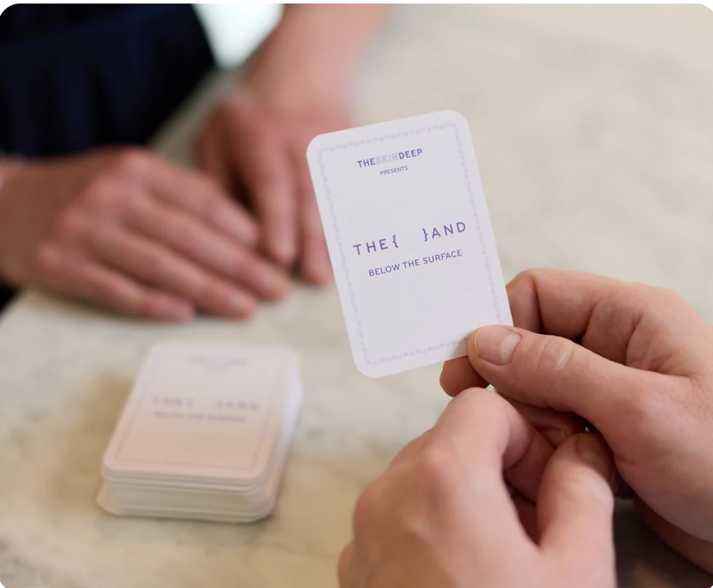 Two people at a table, one handing over a Below the Surface conversation card from The Skin Deep